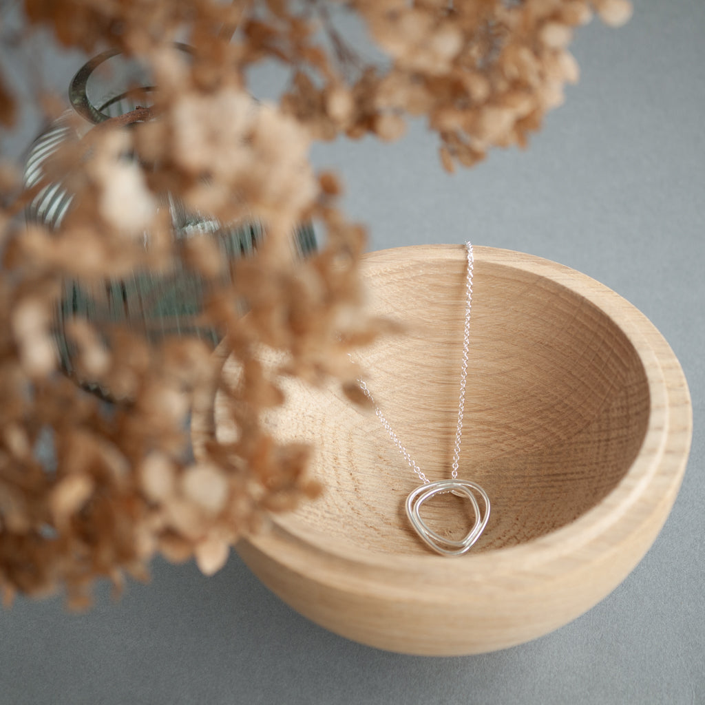 Silver necklace with a triangle pendant in a wooden bowl, surrounded by dried flowers on a gray background